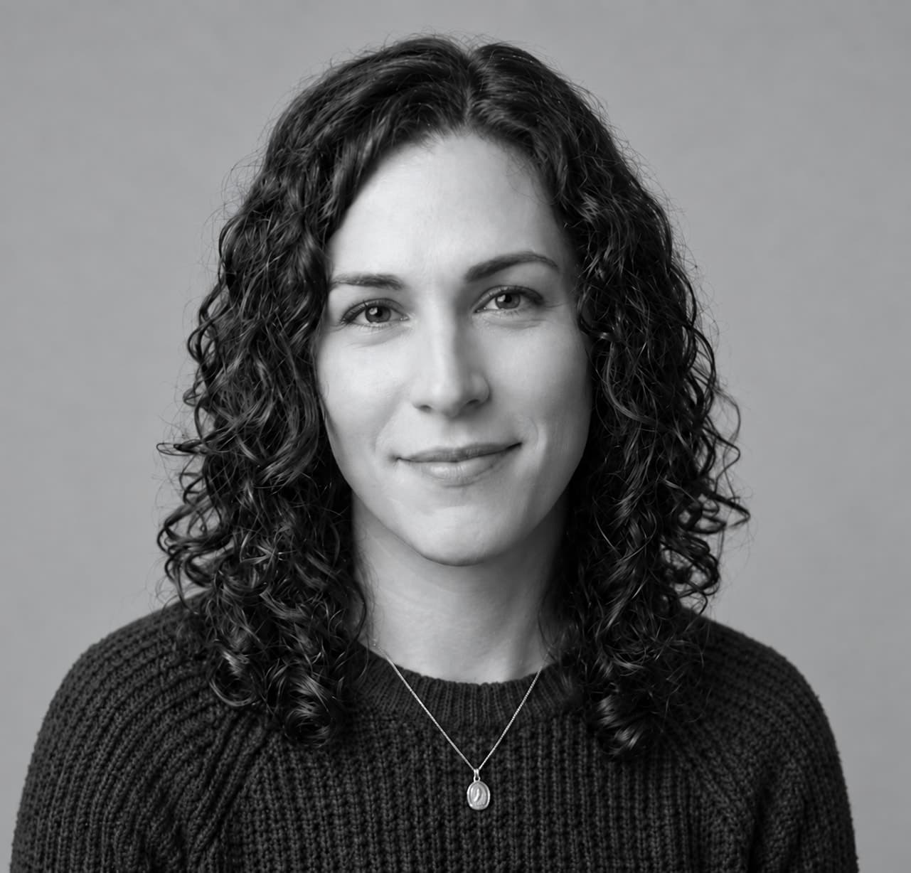 Black-and-white portrait of a woman with dark curly hair in a dark knit sweater, looking at the camera with a slight smile